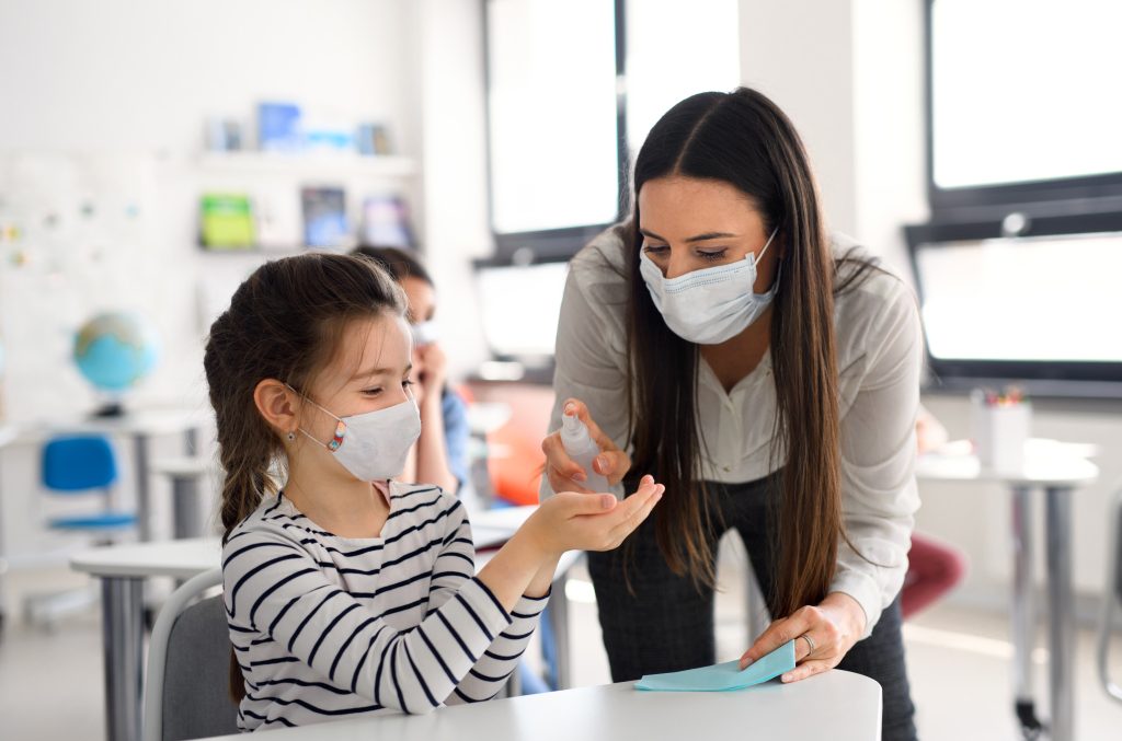 Teacher and children with face mask back at school disinfecting hands