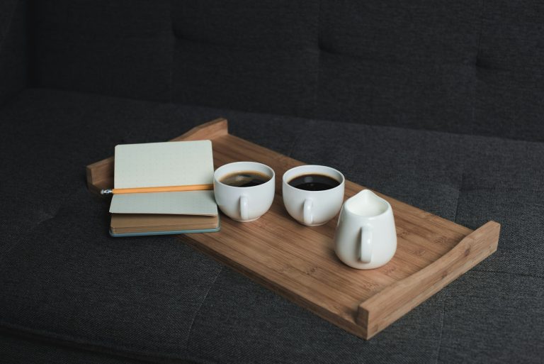 Two cups of coffee, jug of milk and notebook with pencil on wooden tray on sofa