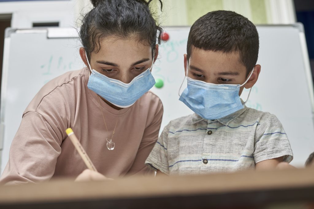 Young female art teacher with a facemask helping a young pupil draw during the class at school